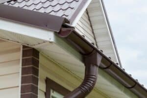 Corner of house with new brown metal tile roof and rain Gutter. Metallic Guttering System, Guttering and Drainage Pipe Exterior